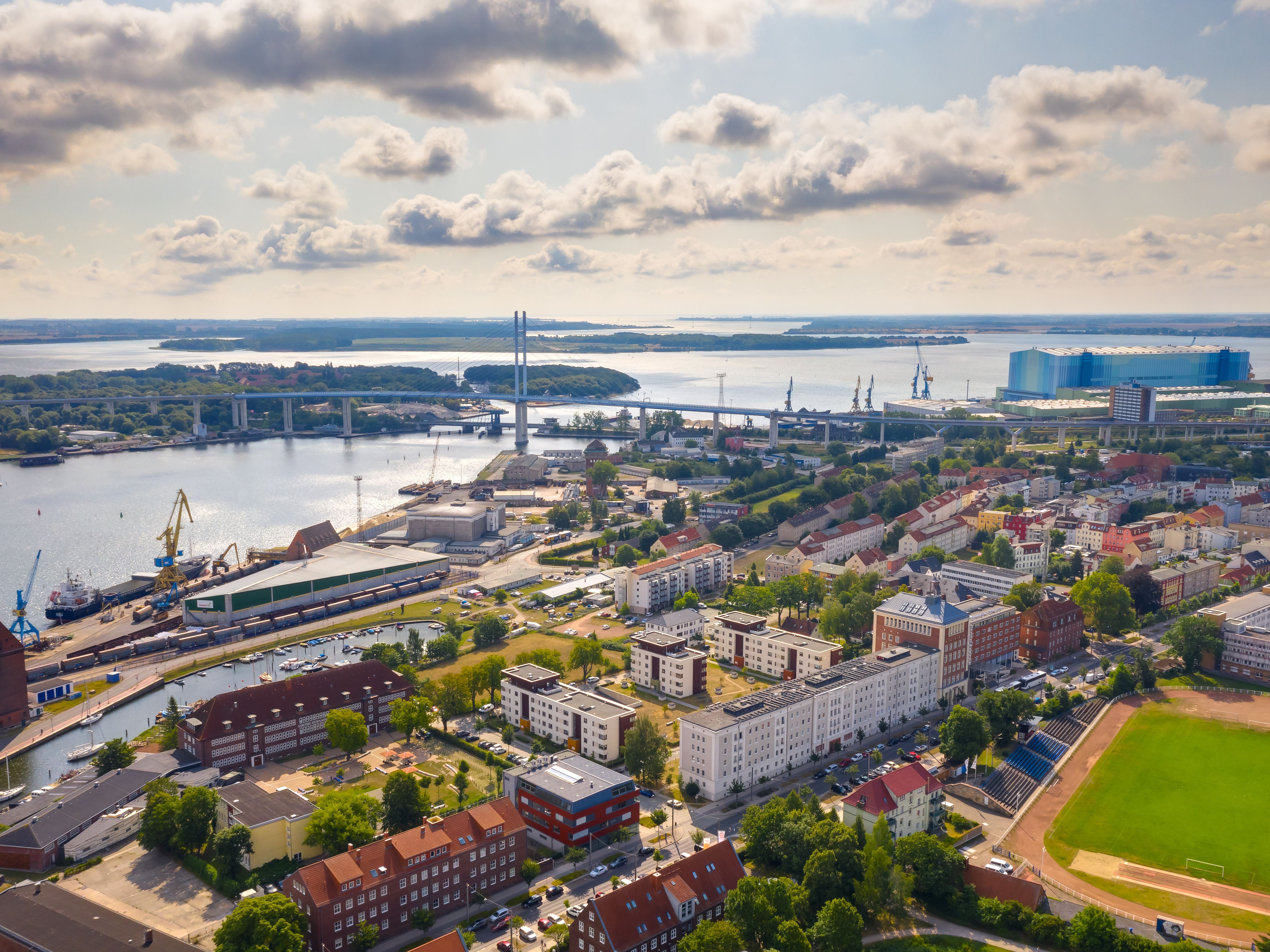 Ein Luftbild zeigt eine Stadt am Wasser, mit einer Brücke, die zwei Ufer verbindet, und industriellen Strukturen im Hintergrund. Die Mischung aus Wohnvierteln, Gewässern und industriellen Anlagen gibt einen Eindruck von der Vielfalt städtischer Umgebungen.